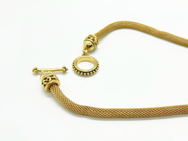 This 1990s pendant necklace features a meticulously crafted design with round purple faceted gemstones set in bezel-mounted antiqued gold metalwork. The ornamental pendant is suspended from a rounded mesh chain, measuring 16" secured with a toggle clasp.