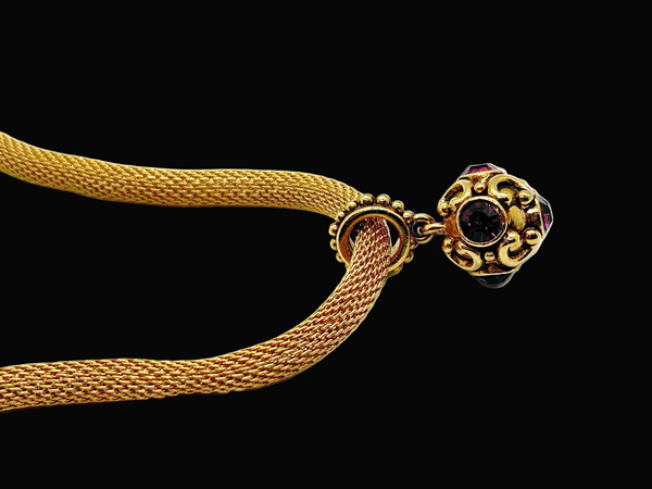 This 1990s pendant necklace features a meticulously crafted design with round purple faceted gemstones set in bezel-mounted antiqued gold metalwork. The ornamental pendant is suspended from a rounded mesh chain, measuring 16" secured with a toggle clasp.
