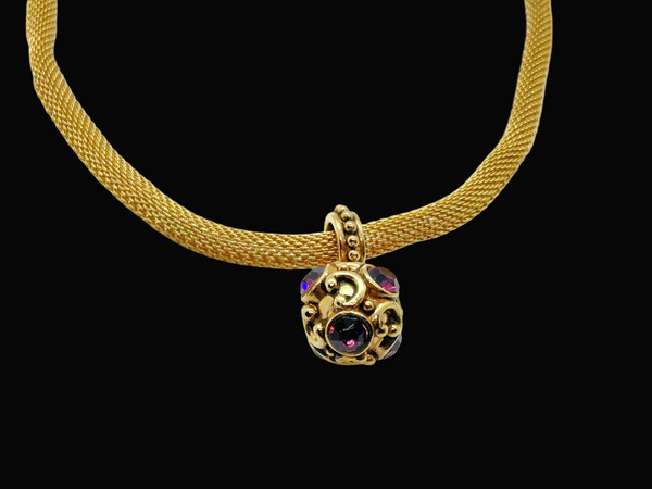 This 1990s pendant necklace features a meticulously crafted design with round purple faceted gemstones set in bezel-mounted antiqued gold metalwork. The ornamental pendant is suspended from a rounded mesh chain, measuring 16" secured with a toggle clasp.
