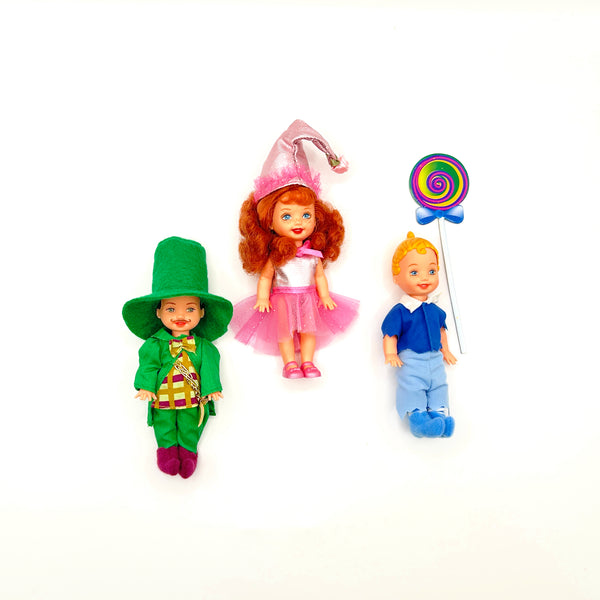 Three loose vintage 1999 Mattel Barbie Kelly Club mini dolls from The Wizard of Oz line arranged on a white background: Tommy as Mayor Munchkin in a green felt top hat and jacket on the left, Kelly as Lullaby Munchkin in a pink conical hat and tutu in the center, and Tommy as Lollipop Munchkin holding a colorful swirl lollipop on the right