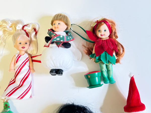 A group photo of three miniature 4-inch Mattel Kelly Club Christmas dolls from the 2001 Holiday Collection. On the left is Peppermint Kelly with blonde hair and red streaks in a striped gown. In the center is Snowman Tommy wearing a green scarf. On the right is red-haired Poinsettia Jenny next to her green plastic pot accessory, with a small fabric Santa hat off to the side