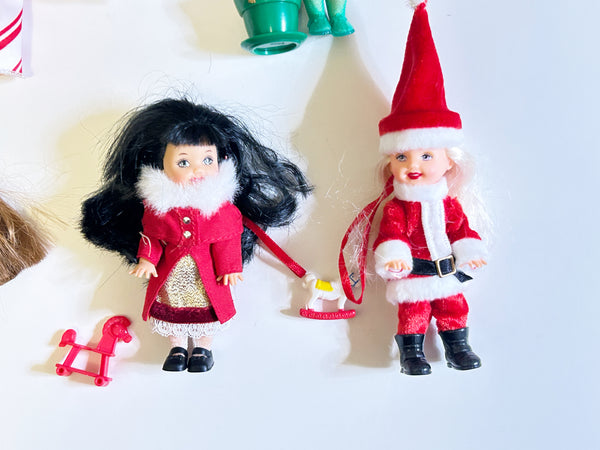 Product listing image featuring two miniature Mattel Barbie Kelly Club holiday dolls. On the left is the 2001 Caroling Lorena doll with black hair, wearing a white-trimmed red coat. On the right is the 2003 Santa Claus Kelly doll with blonde hair, wearing a red Santa hat and suit. Small holiday toy accessories sit on the white background