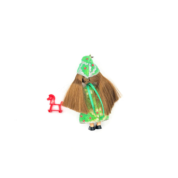 Back view of a vintage miniature Christmas tree ornament doll featuring long, straight brown hair trailing past her waist. She is wearing a green holiday dress with red and white floral patterns, a matching cone hat with white trim, and tiny black shoes. A small red plastic rocking horse accessory sits next to her against a solid white background
